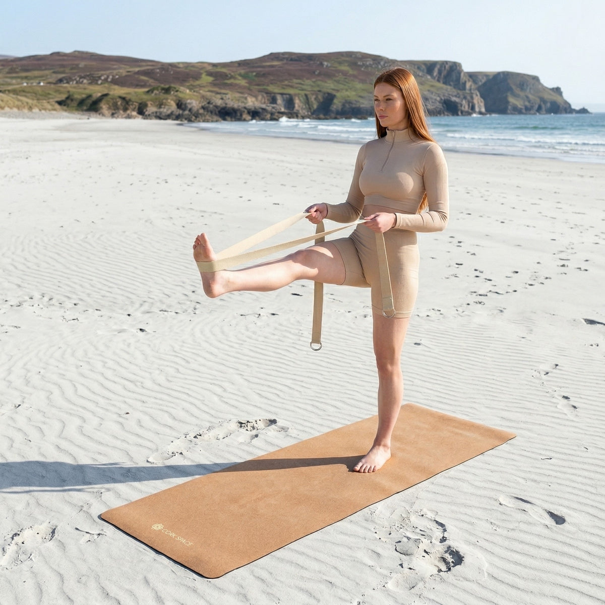 Woman balancing on a cork yoga mat on a sandy beach, using a strap to support her extended leg.